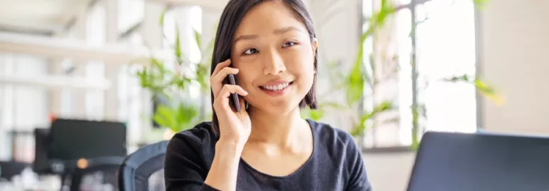 female Chinese white-collar professional in her 30s answering her mobile phone while sitting in front of a laptop in an office setting.